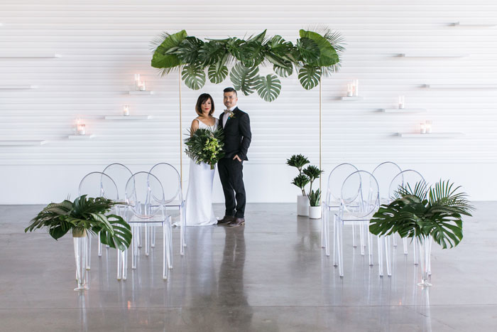 Modern, minimal, tropical wedding ceremony arch with monstera, banana leaves, elephant ear, and ghost chairs.