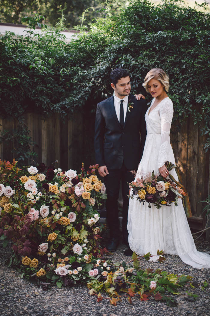 Boho couple pose next to rustic wheel barrow overflowing with fall flowers in shades of blush, gold, and berry