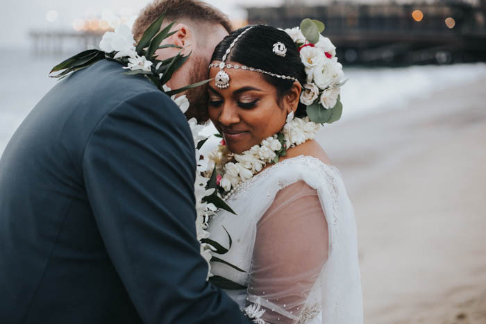 The cutest couple share an intimate moment on the beach for a private lei exchange featuring orchids, ti leaf, & tuberose following their wedding ceremony. The bride wears an ornate yet modern take on a Sri Lankan floral headpiece featuring ranunculus, spray roses, and eucalyptus.