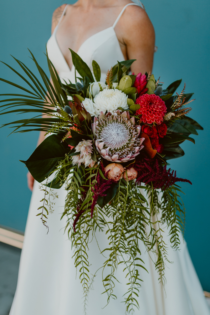 A portrait of Bellamy's large tropical bridal bouquet which features protea, dahlias, garden roses, and blushing bride protea as well as tropical greenery, monstera and pepperberry in shades of blush, coral, red, and white. 