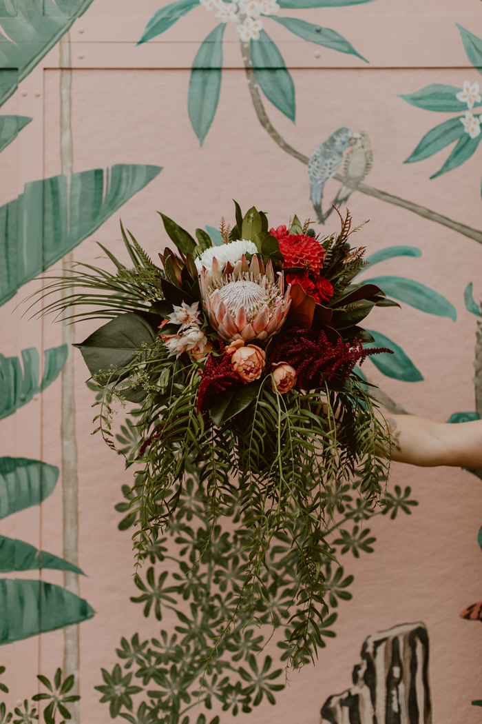 A modern tropical cascade bouquet featuring king protea, peach garden roses, red dahlias, and tropical greenery like monstera, areca palm and more. 
