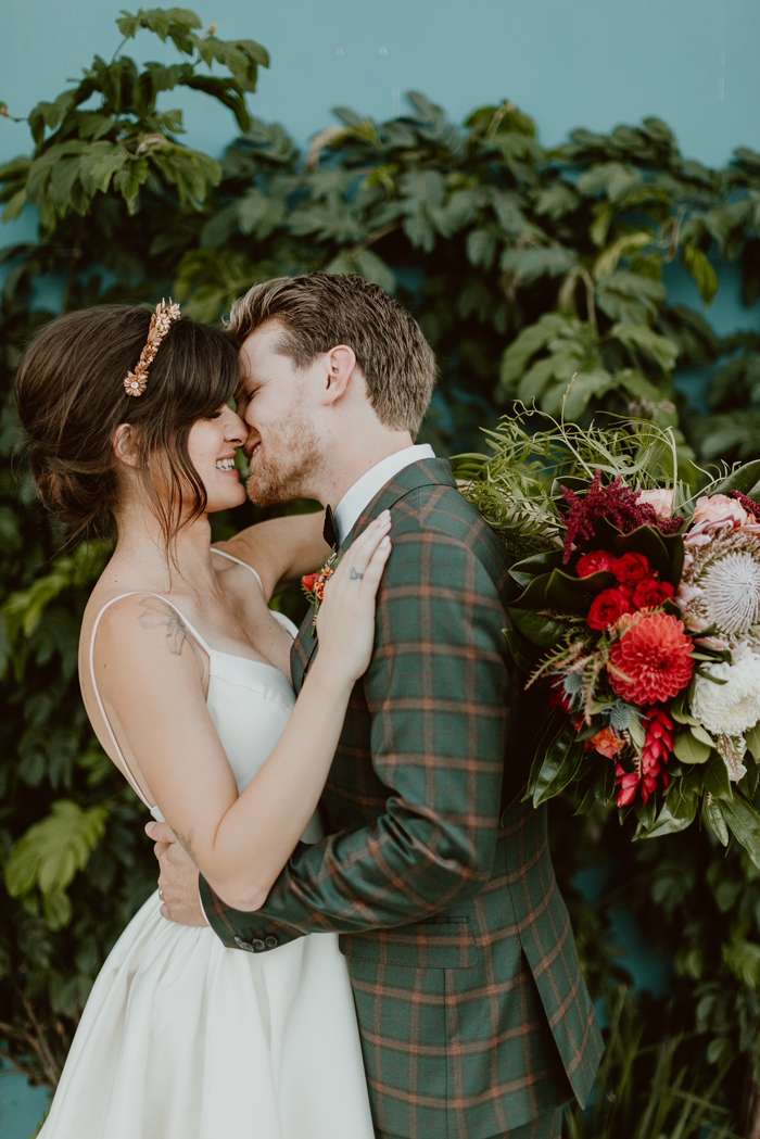 The stylish couple shares a sweet kiss at their romantic and tropical wedding at Valentine in DTLA. She holds a bold tropical bouquet and he wears a stylish tartan suit.