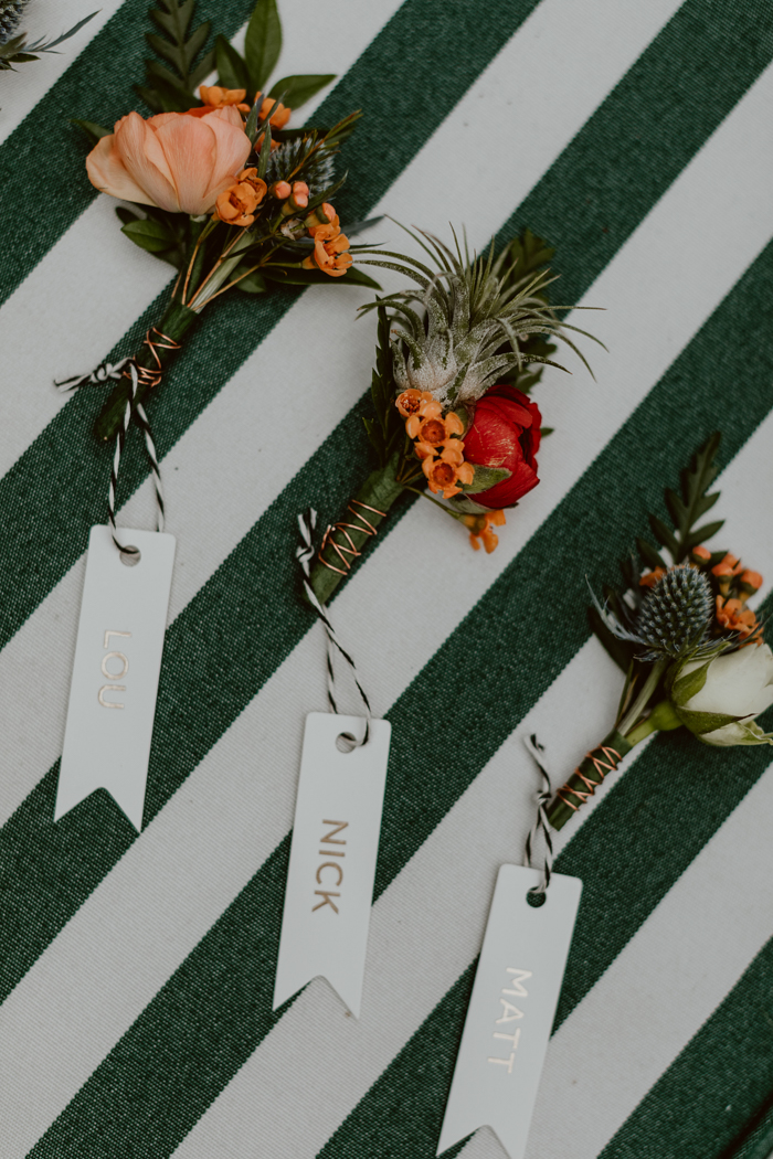 Tropical boutonnieres with custom name tags feature coral and red ranunculus, with accents of thistle, tiny tropical air plant, and wax.