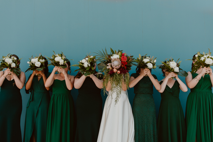The Bride and bridesmaids pose for a wedding portrait with their tropical bouquets held in front of their faces- the bridesmaids wear dresses in shades of green and they all stand against a teal wall.