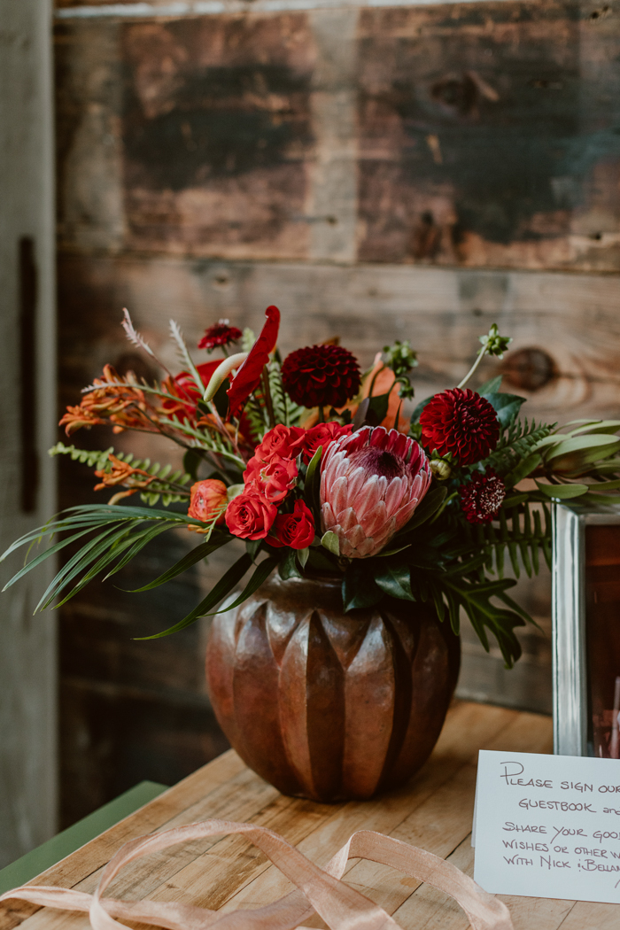 A bold welcome arrangement featuring protea, red dahlias, anthurium, kangaroo paw and other tropicals in a vintage copper vase.