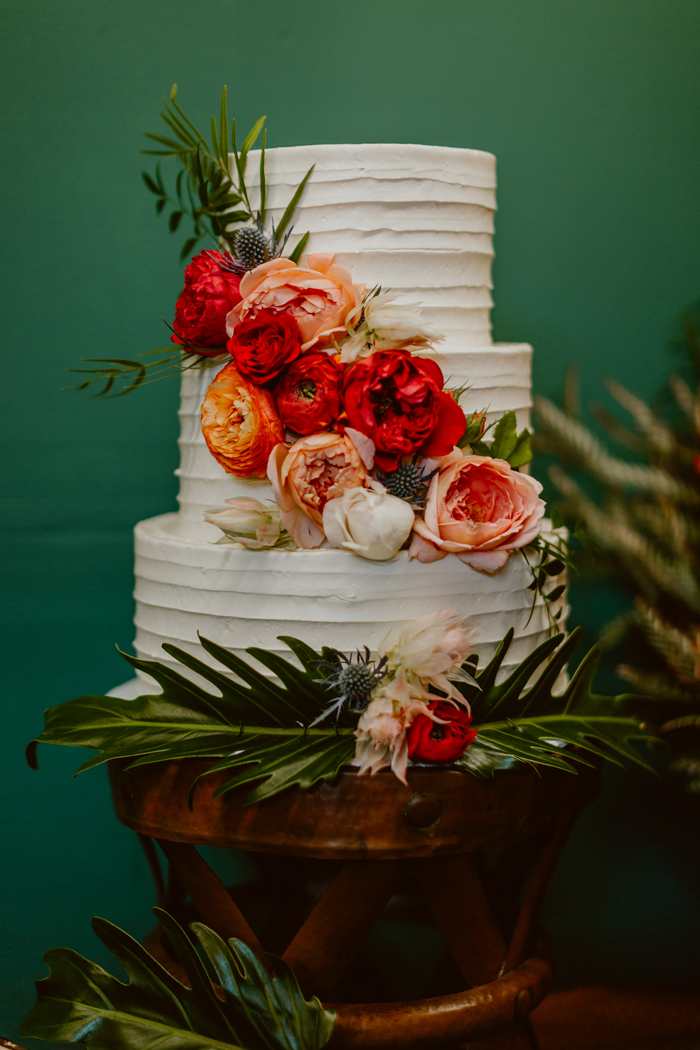 The 3 tier wedding cake is decorated with ranunuculus, garden roses, blushing bride protea and tropical greenery in shades of red, coral, orange, peach and blush. tropical greenery and bold red and coral flowers