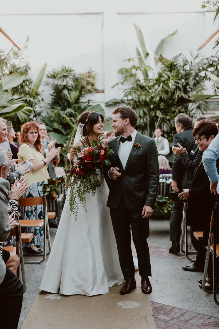 The stylish couple recess up the aisle after their ceremony- she carries a modern tropical cascade bouquet and he wears a tartan suit with a bold red boutonniere. 