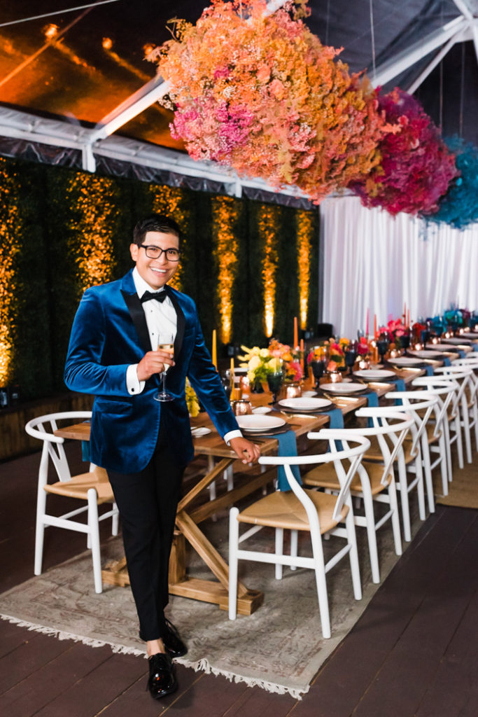 The guest of honor wears a luxe blue velvet dinner jacket and is standing under 21 feet of bold and colorful floral clouds.