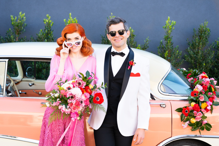 A colorful Valentine's Day wedding couple pose with their vintage pink car.