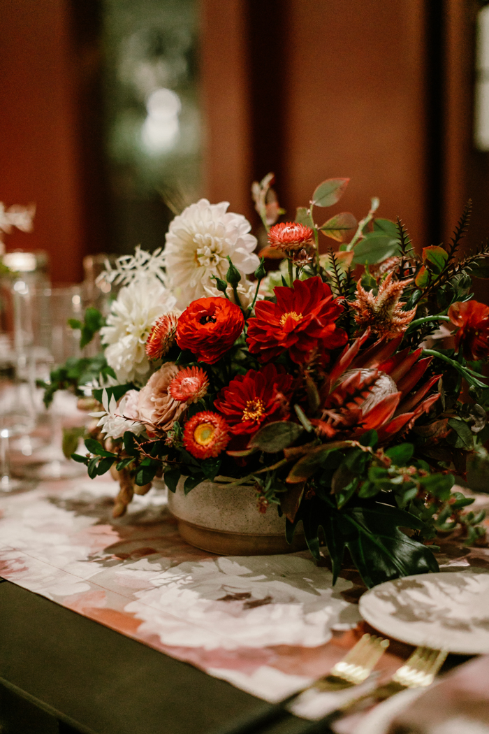 Lush centerpiece featuring King Protea, Ranunculus, Strawflowers, Dahlias, and more in shades of red, mauve, and white. 