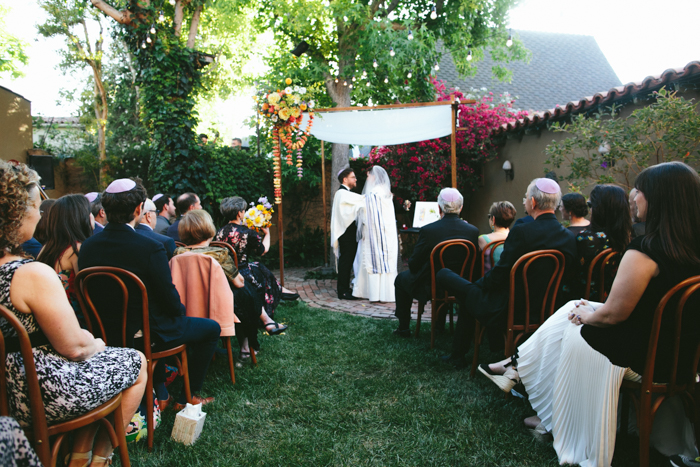 Our couple stands under the Chuppah, adorned in local spring flowers in shades of orange and lavender for their wedding ceremony. 