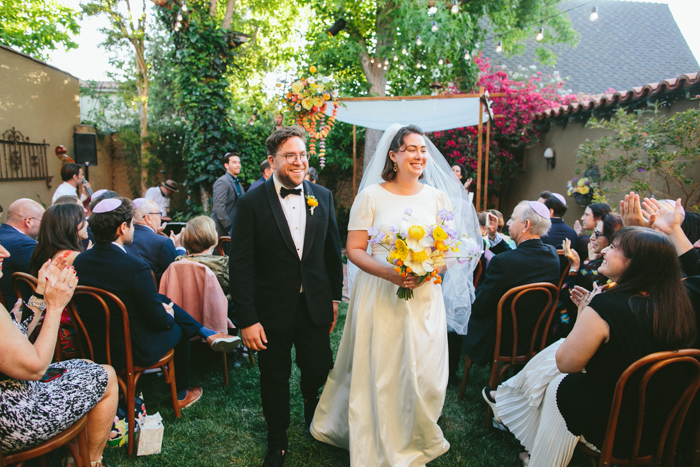 Our couple recesses following their ceremony- our bride carries her airy statement bouquet and the whimsically decorated Chuppah is in the background. 