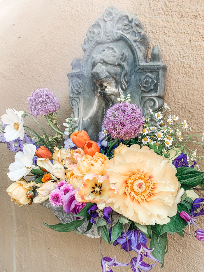 A site specific floral installation by Winston & Main in an existing fountain features locally sourced Cosmos, Poppies, Tulips, and Allium in shades of lavender, orange, & yellow.