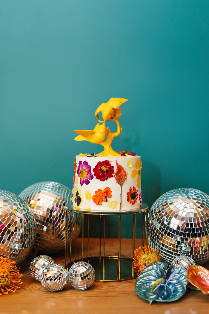 An incredible fresh-pressed flower cake by Nicole Bakes Cakes, styled with a yellow bird cake topper, disco balls &  bright tropical blooms.