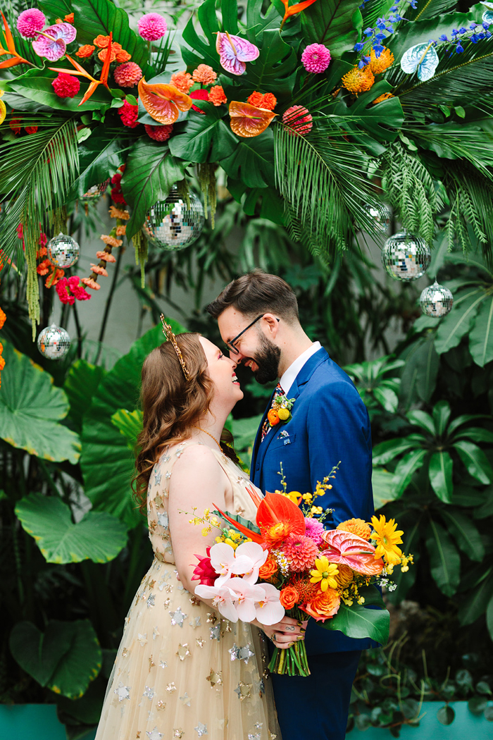 Our fun couple share a first kiss under our bold, colorful, & tropical hanging floral installation. 