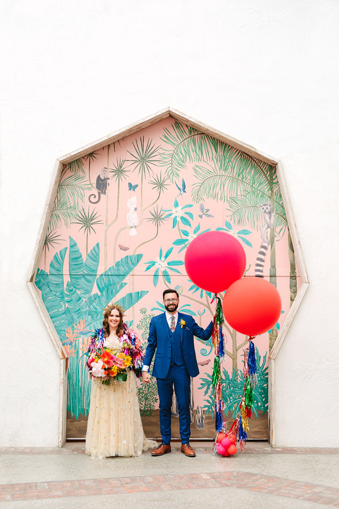 The cutest quintessential wedding portrait at Valentine- the bride wears a fab fringe jacket and holds a colorful tropical bouquet and the groom holds giant colorful balloons.