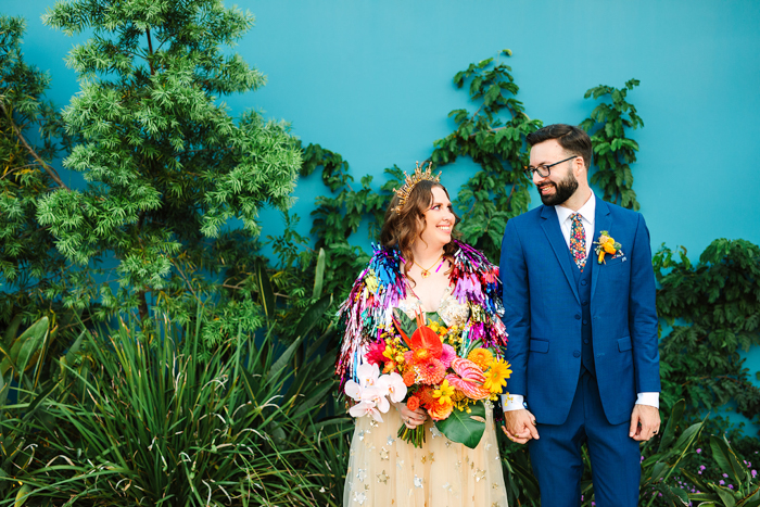 A cute wedding portrait at Valentine DTLA, featuring a bold fringe jacket by Rachel Burke, a teal wall, our colorful bouquet and our cute couple.
