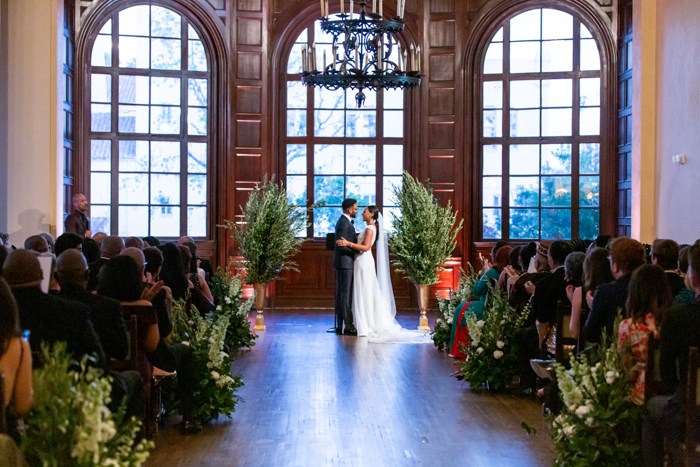 A wide view of the ceremony from back of the room. Our couple holding and facing each other at the altar and surrounded by nature-inspired lush greenery and white flowers.