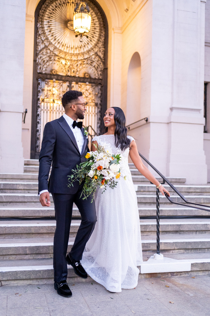 A wedding portrait of the couple on the steps of the historic Ebell of Los Angeles, featuring a modern cascade bouquet in fresh white & green with orange and yellow accents.