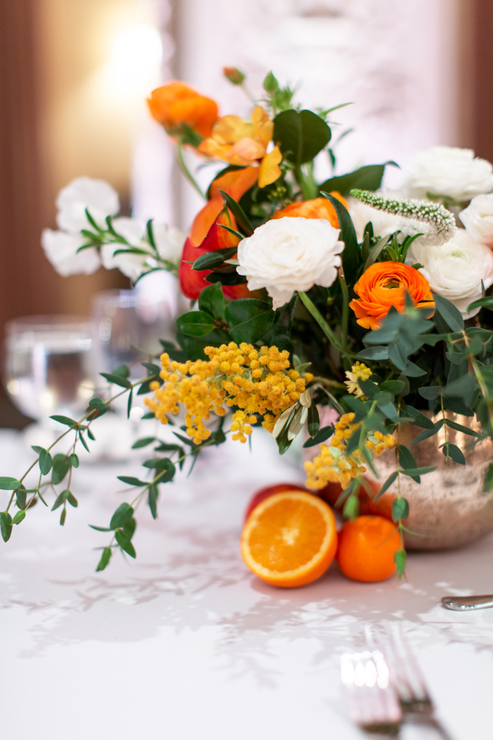 A bright & textural centerpiece features lush spring blooms in shades of white, orange and yellow with mimosa and citrus accents on the table.