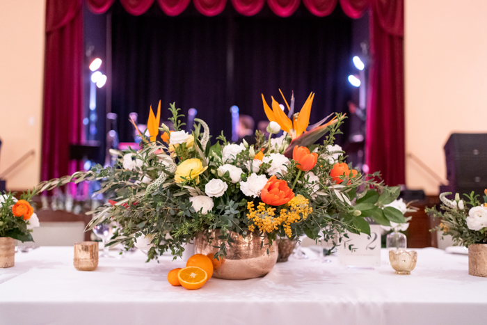 A bold centerpiece at the head table in front of the wedding reception room's stage, features dramatic Birds of Paradise and full white, orange and yellow flowers.