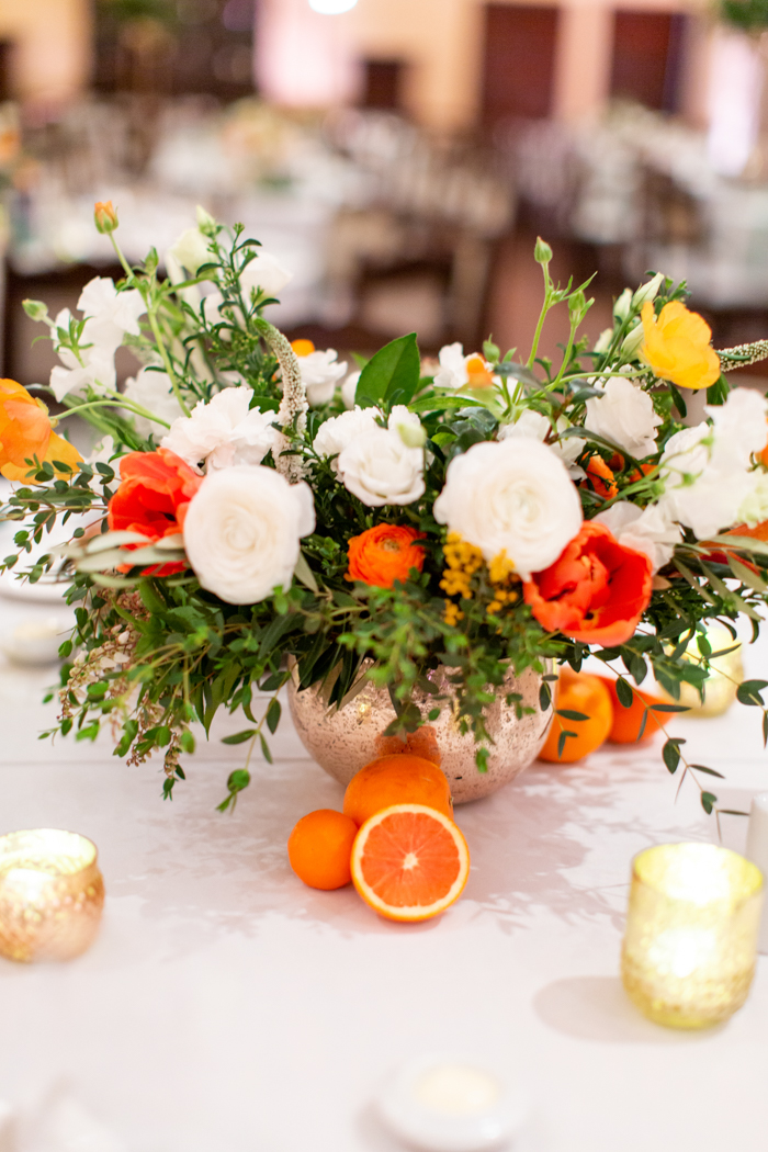 A cheerful centerpiece with white flowers and citrus-colored accent blooms in a gold bowl. Whole & halved oranges were artistic touches placed next to the bright arrangement with votive candles.