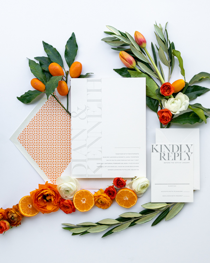 An artistic floral flat lay with ranunculus, olive leaves, and citrus accents for a modern orange wedding.