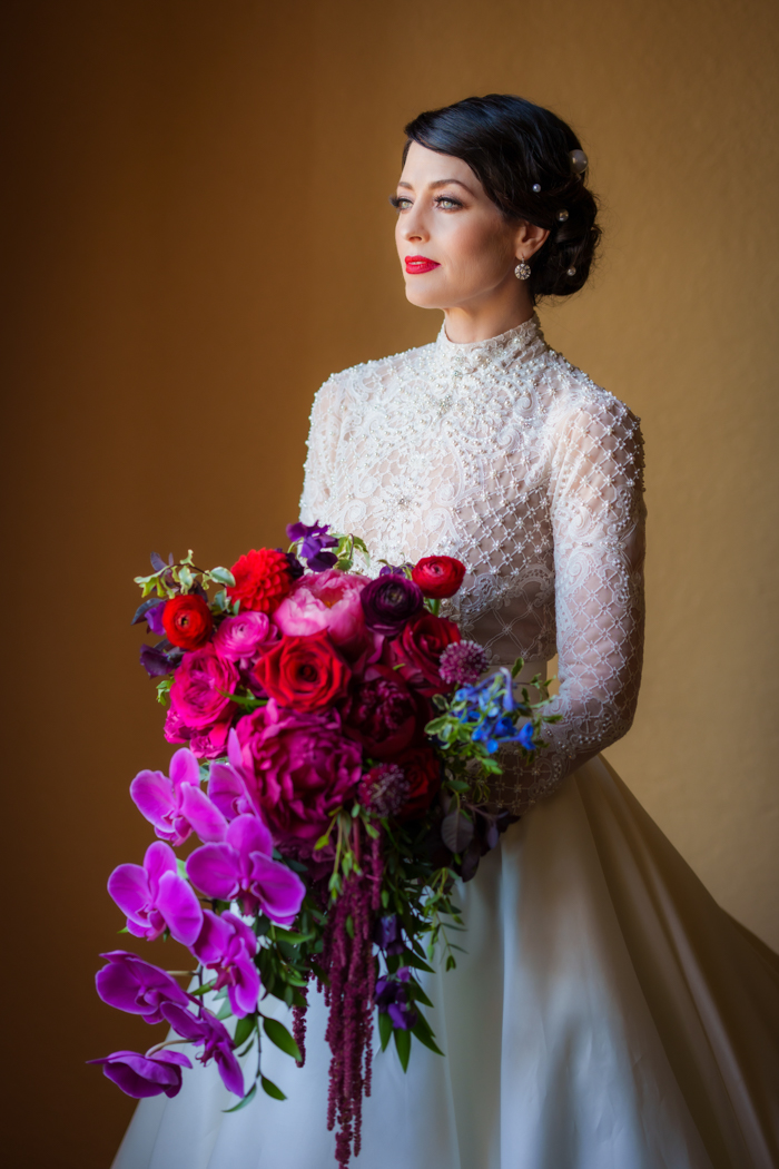 A stunning portrait of the bride in front of an amber wall as she holds her modern cascade bouquet full of orchids, peonies, and garden roses, in shades of pink and red by Los Angeles florist Winston & Main.