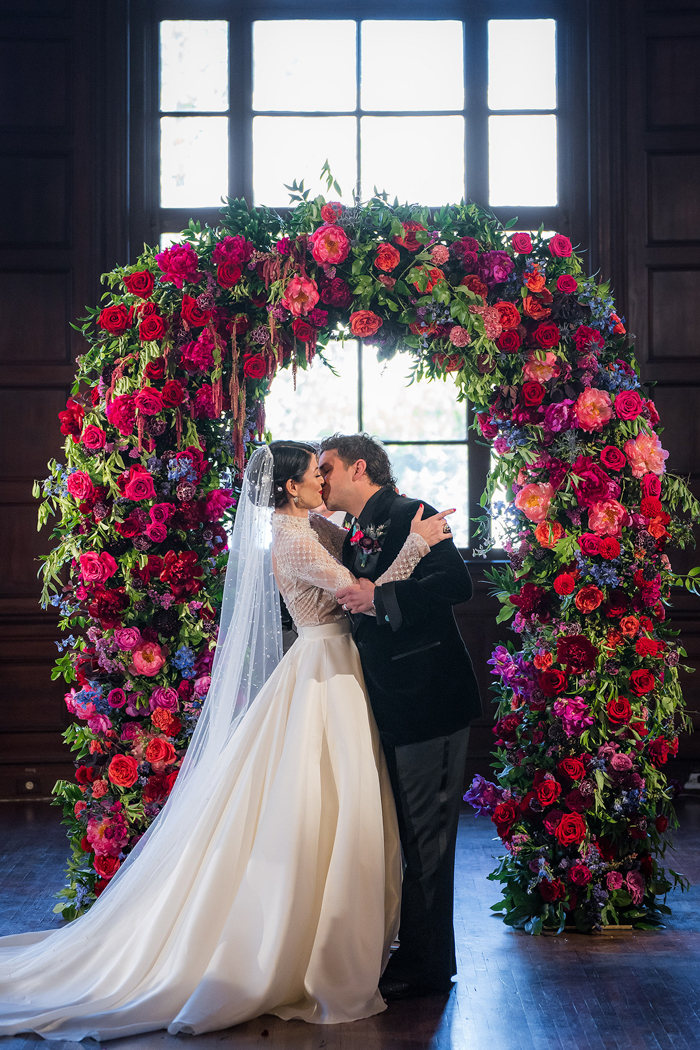 The couple shares their first kiss under a bold and lush wedding arch full of peonies & garden roses, created by Los Angeles florist Winston & Main.