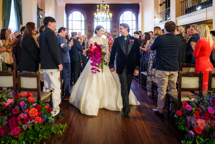 The couple smiling at each other as they walk back down the aisle after their wedding ceremony surrounded by large, colorful aisle arrangements.