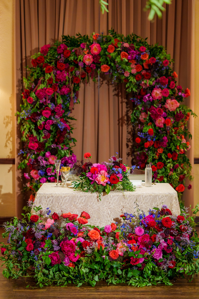 An image of a sweetheart table, with the freestanding ceremony arch, dripping with peonies, orchids, and garden roses, repurposed behind the table.
