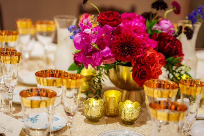 A bright and textured centerpiece with pink orchids and red dahlias in a gold compote, complements gold rimmed wine glasses and votives.