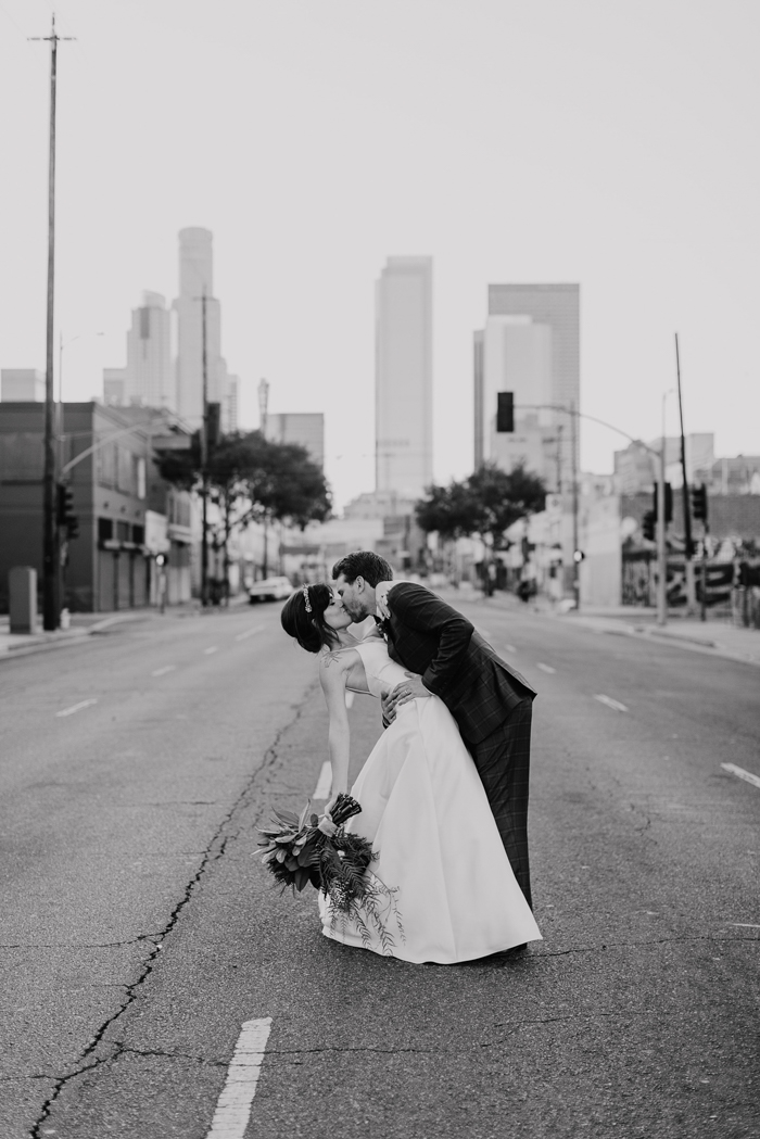 A black & white photo of a couple kissing in the middle of the street with views of DTLA behind them. She holds a large modern tropical cascade bouquet.