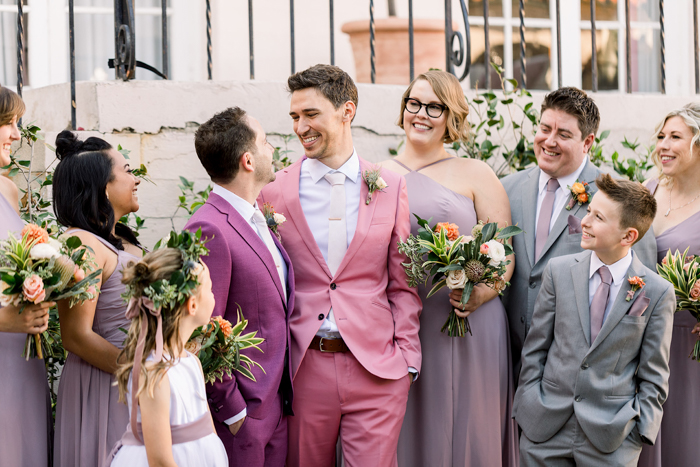 Two grooms in colorful custom suits are surrounded by their wedding party- everyone wears or carries flowers from Los Angeles Floral Designer, Winston & Main.
