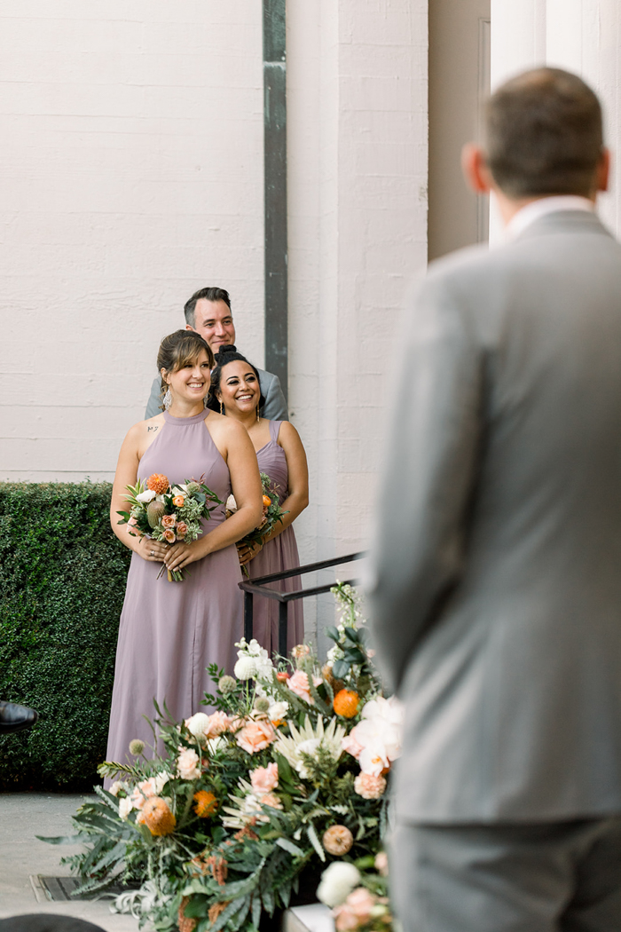 The wedding party smile and laugh during this beautiful same-sex wedding at The Ebell of Los Angeles. They hold textural and organic bouquets in shades of blush, peach, coral and orange by Winston & Main.