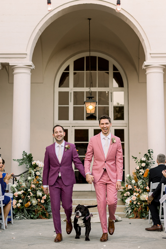 Two grooms in custom, colorful suits walk hand-in-hand after their California wedding ceremony with their dog Black Jack. Behind them is a large-scale floral installation on either side of the ceremony area featuring King Protea, Banksia, Air Plants, Orchids, Dahlias, Scabiosa, Amaranthus, Protea, Rose and more! 