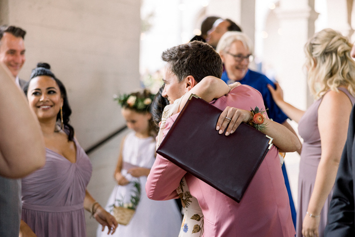 Family and friends exchange hugs, kisses, and smiles with one another celebrating our couple following their ceremony. They wear floral crowns, hold flower baskets, and wear a signature Winston & Main corsage on a brass cuff in wedding colors of blush, peach, and coral. 