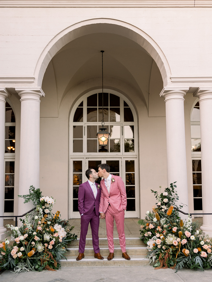Our couple share a kiss in front of their large scale floral installation at The Ebell of Los Angeles full of citrus-toned and sunset-inspired flowers like King Protea, Banksia, Air Plants, Orchids, Dahlias, Scabiosa, Amaranthus, and Rose! 