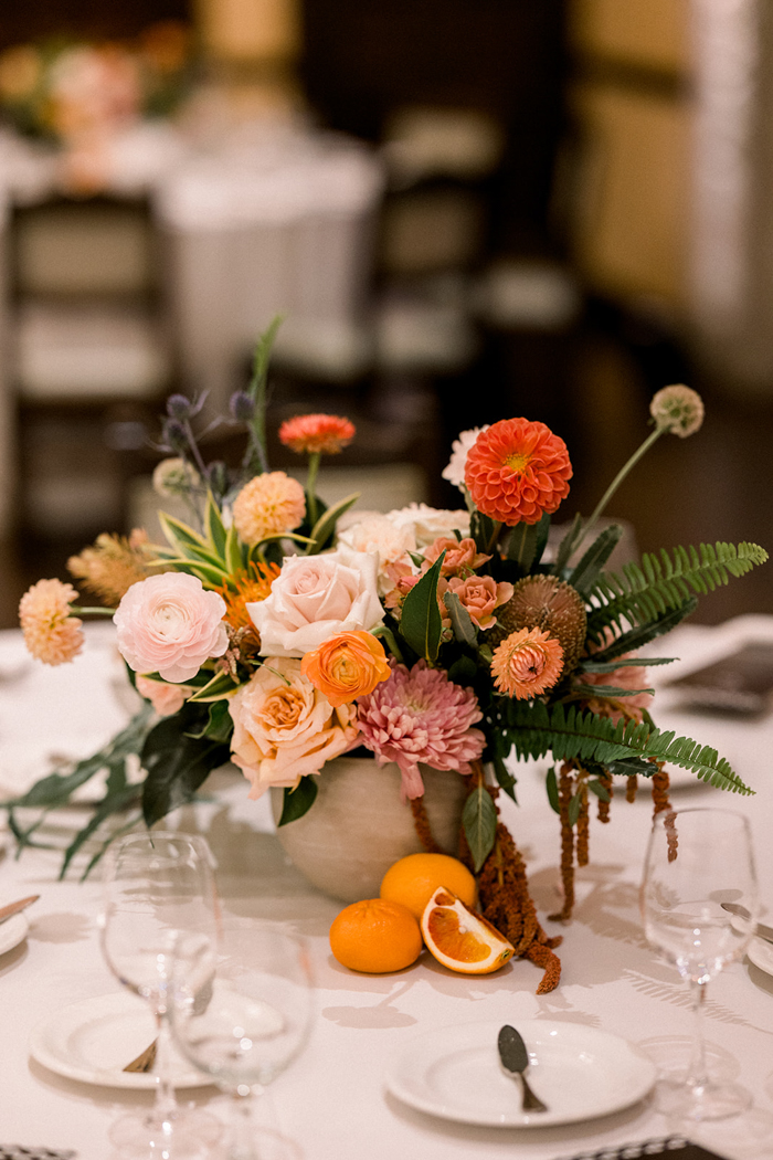 Reception tables feature lush and textural floral centerpieces full of orange Dahlias, blush Garden Roses, Ranunculus, Scabiosa, Strawflower, and Amaranthus, and are styled with fresh fruit for this stunning same-sex California wedding. 