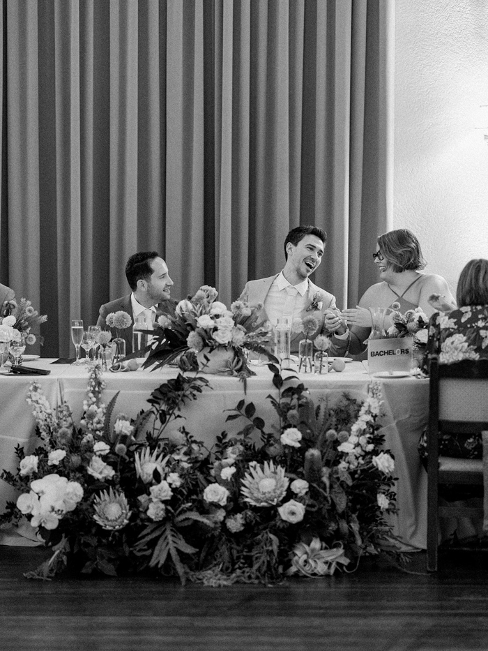 Our happy couple exchanges smile and laugh with family and friends during their California wedding reception at The Ebell of Los Angeles. Their head table is adorned with a large scale floral installation by Winston & Main that is textural, tropical, and organic. 