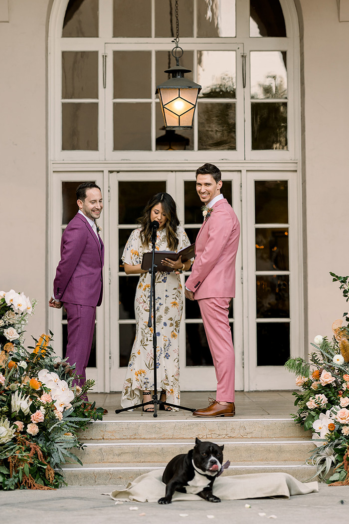 A wedding ceremony on the steps of The Ebell of Los Angeles- the two grooms wear colorful suits and are surrounded by a large scale floral installation while their dog relaxes on his dog bed in front of them. 