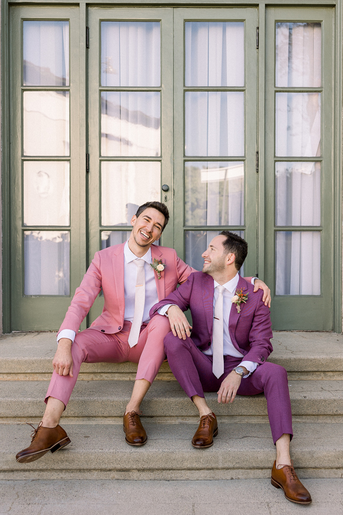 Our happy couple sits on steps wearing custom pink and purple wedding suits for their California Wedding at The Ebell of Los Angeles.